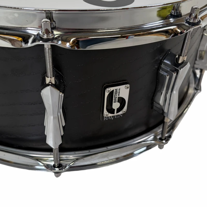 Black snare drum with chrome tension rods and badge, close-up shot highlighting drum details.