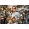 Female drummer holding cymbal with drum kit and percussion instruments in the background.