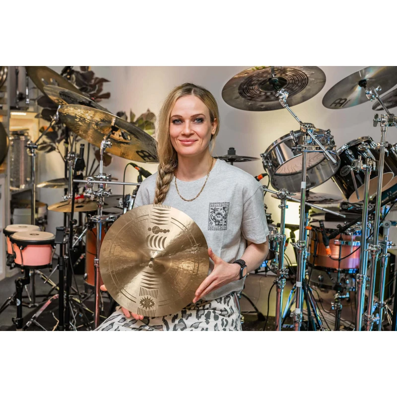 Female drummer holding cymbal with drum kit and percussion instruments in the background.