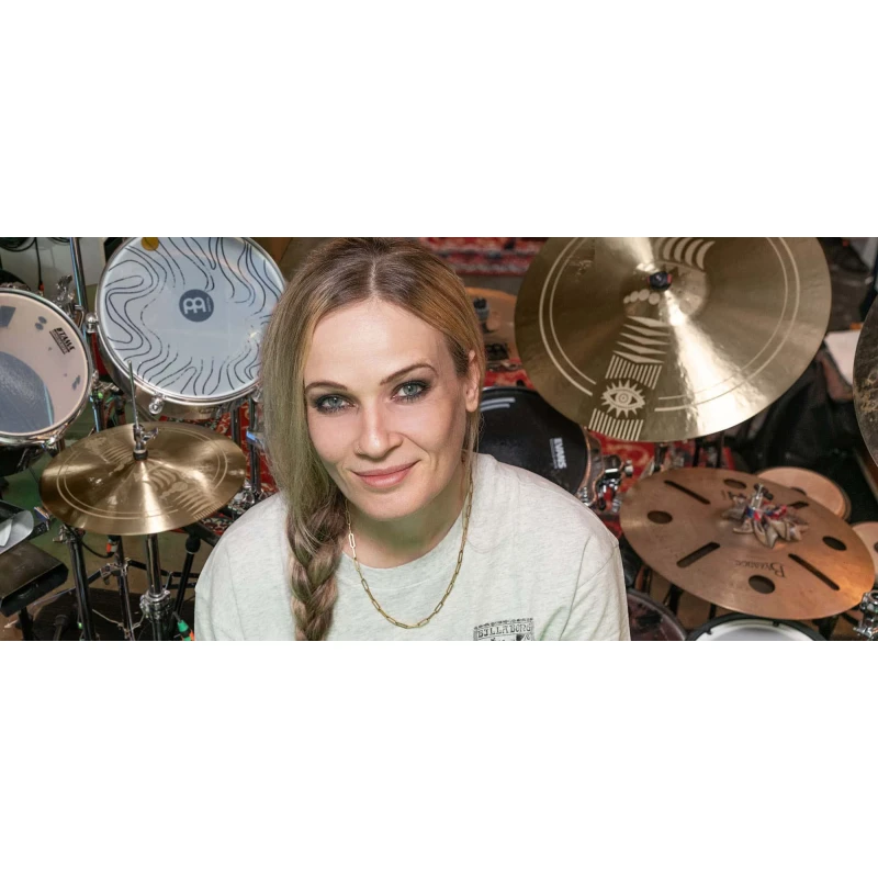 Female drummer smiling with drum kit and cymbals in a music studio.