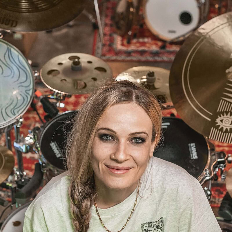 Female drummer smiling among various cymbals and drums in a music studio.